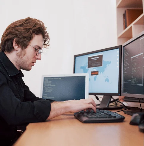 Ein Mann tippt auf einer Computertastatur, vor ihm stehen ein Laptop und PC-Monitore.